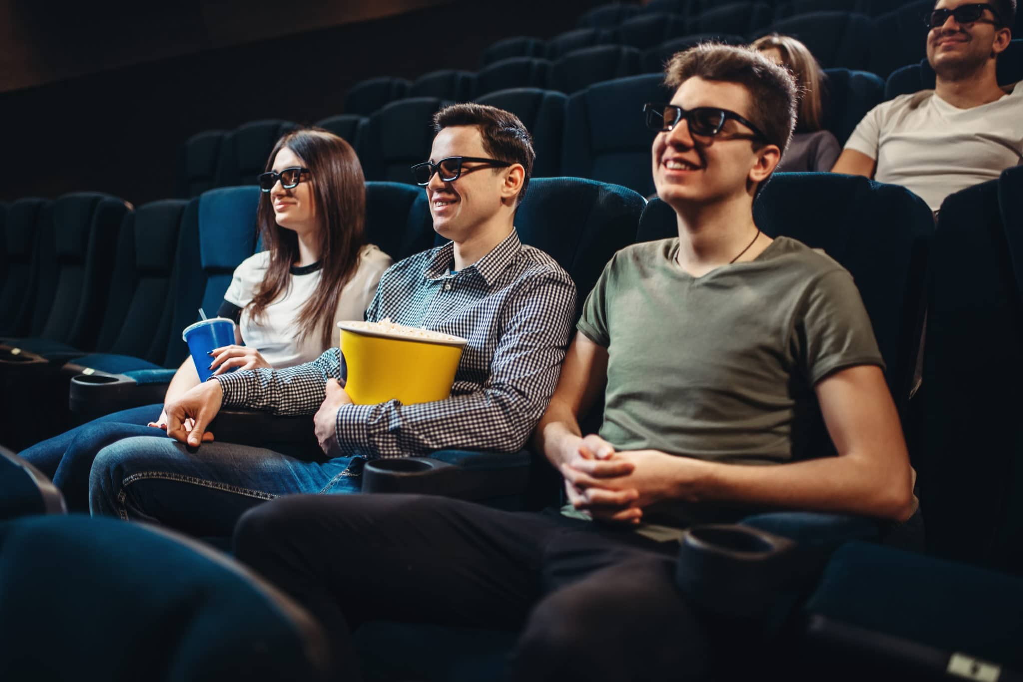 3 young people watching movie with popcorn wearing 3d glasses in a theater like AMC.